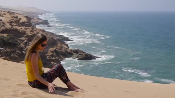 Jovem mulher sentada na areia e caindo sandfrom mão com mar oceano rochas no fundo — Vídeo de Stock