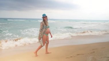 Swimsuite ve kumlu bir plaj dalgalar deniz boyunca yürürken tunik şık hamile kadın.
