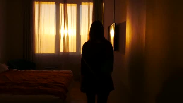 Silueta de una chica que va hacia la ventana y desvela cortinas. Impresionante luz del sol de oro — Vídeos de Stock