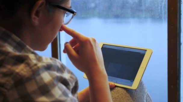 Mujer caucásica en gafas sentadas junto a la ventana y utilizando la aplicación de mapa en su tableta — Vídeos de Stock