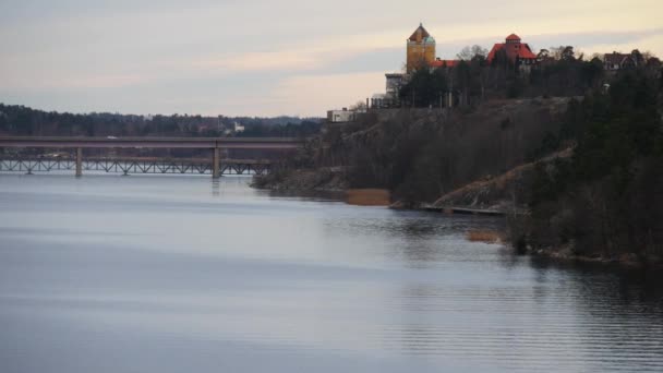 Скалистый берег реки с красивым зданием и мостом через реку — стоковое видео