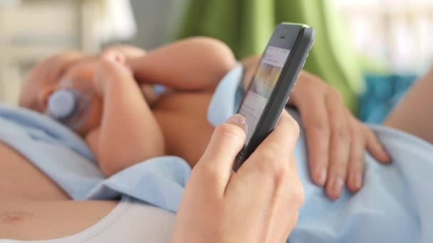 Donna che tiene il neonato su un petto e guarda le foto da internet con il cellulare — Video Stock