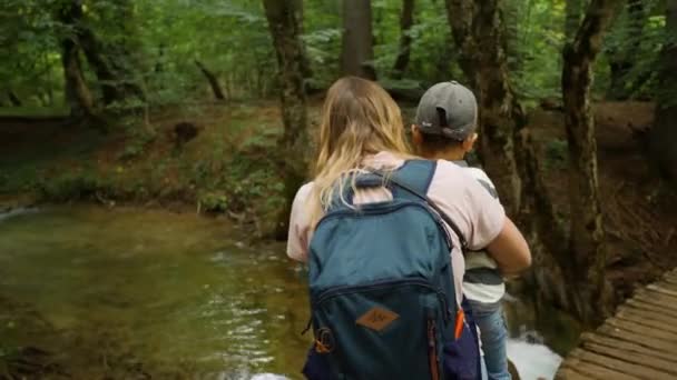 De vrouw en haar kind kijken op een heldere stroom. Familie wandelingen in een groen bos — Stockvideo