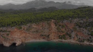 Dik yamacın eteğinde küçük bir sahil. Deniz sularının üzerindeki yeşil orman. Bulutlu bir gün