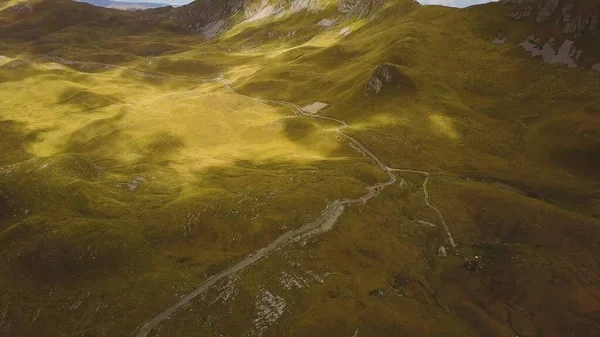 Dağ yolu çimenli tepeler ve kayalar arasında dönüyor. Bulutların gölgeleri ve yukarıdan gelen güneş ışığı