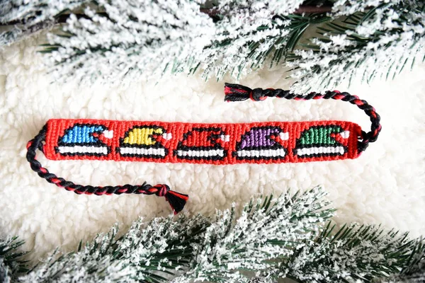 Woven friendship bracelet with bright alpha pattern Multicolored christmas hats, handmade of thread, next to snowy fir branches.DIY Christmas or New Year gift idea