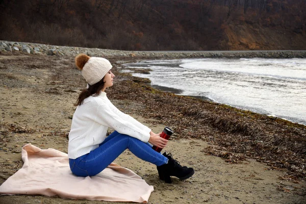 Young woman in white sweater, hat with pom-pom, sitting on the shore of the Japanese sea with closed eyes, inhaling the aroma of winter air. She holding thermos with tea. Copy space