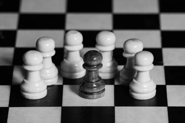 Chess wooden pieces on the board. A black pawn on the playing field surrounded by white pawns. Management or leadership. The concept of race equality. Close-up, selective focus. Black and white photo — Stock Photo, Image