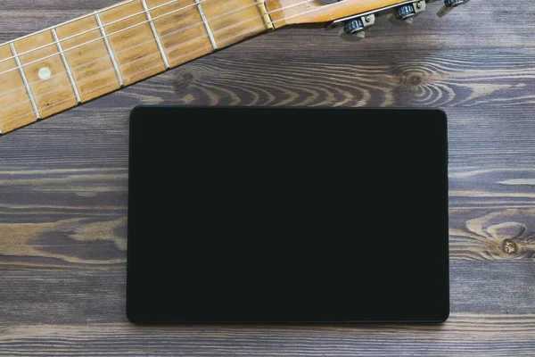 Guitar neck and black tablet on a brown wooden background. Close-up, copy space, mock up. The concept of online guitar courses.