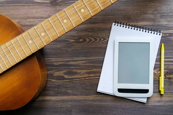 Online education. Part of the guitar, a notebook and part of the tablet, a yellow pencil lie on a wooden background. Top view, close-up. Mock up for advertising guitar courses.