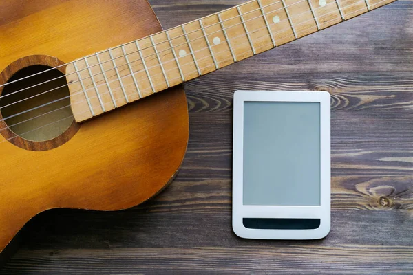 A piece of guitar and a white tablet vertically placed next to it. Brown wooden background, copy space, layout. The concept of advertising online guitar courses.