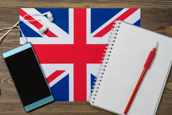 A phone, headphones, spiral notebook, and red pencil rest on the flag of the United Kingdom. Brown wooden background, top view. English language course online . Layout for advertising