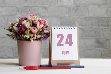 24 Temmuz. Ayın 24 'ü, takvim randevusu. Pembe bir vazoda bir buket çiçek, iki kalem ve tahta bir yüzey üzerinde günü olan bir takvim..