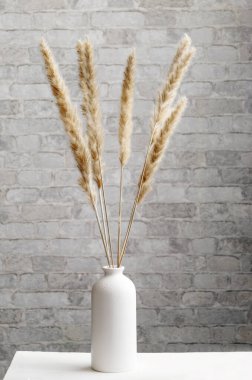 Bouquet of brown dried spikelets of grass in a white vase against a brick wall. Close-up, vertical image, minimalistic interior.