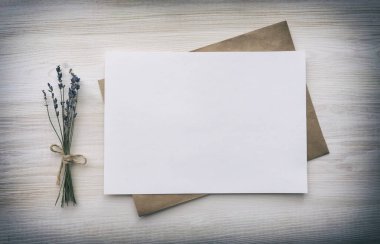White blank sheet of paper with a brown envelope and a dry bouquet of lavender flowers on a white wooden background. Top view, copy space, mockup.