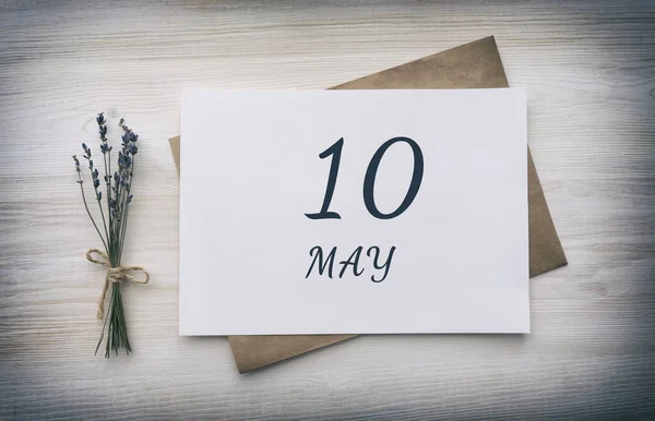 10 Mayıs. Ayın onuncu günü, takvim tarihi. Kahverengi zarflı beyaz bir kağıt, ahşap arka planda bir demet kuru lavanta çiçeği. Bahar ayı, yılın günü konsepti..