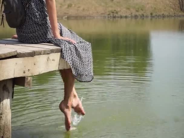 Dívka sedí na dřevěném mostě, na molu, houpe bosýma nohama ve vodě — Stock video