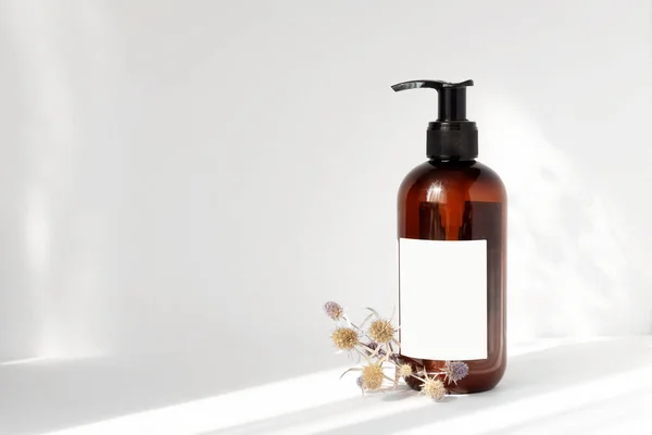 Brown bottle with dispenser, empty label and bouquet of dried flowers next to it on white background in rays of light, with falling shadow. Concept of natural cosmetics.