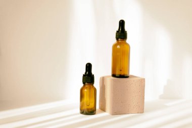 Two bottles, made of brown glass, with dropper lids on light background, in rays of sunlight. One bottle stands on square stand, second next to it. Concept of beauty industry.