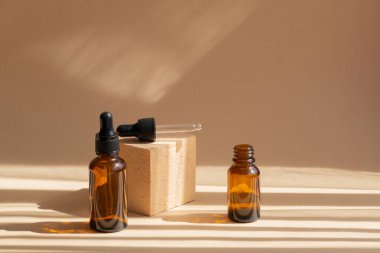 Two glass bubbles on brown background, in rays of sunlight, cast shadow on the surface. One bottle with black cap, cap-pipette from second, lies on square stand. Concept of the beauty industry.