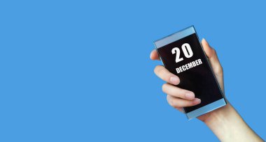 20 Aralık. Ayın 20. günü, takvim tarihi. Kadının elinde mavi izole arka planda boş ekranlı cep telefonu var. Kış ayı, yılın günü konsepti..