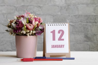 12 Ocak. Ayın 12. günü, takvim randevusu. Pembe bir vazoda bir buket çiçek, iki kalem ve tahta bir yüzey üzerinde günü olan bir takvim...