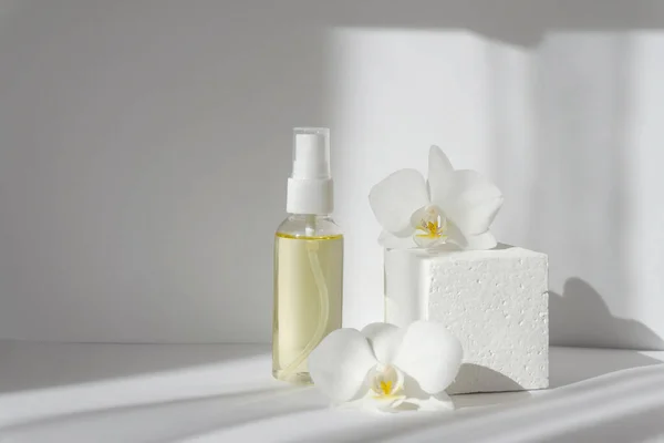 Mock-up of glass bottle with spray gun on white background in morning rays of light. Improvised 3d podium for product demonstrations, surrounded by orchid flowers. Concept of skin care, spa treatments