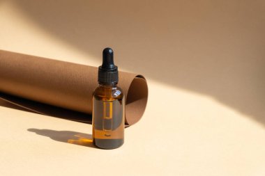 Yuvarlanmış kahverengi kağıt rulosunun arka planında cam damlatan şişenin maketi. Nesne güneş ışınlarından sert gölgeler bırakır. Vücut ve yüz bakımı kavramı, doğal kozmetik..