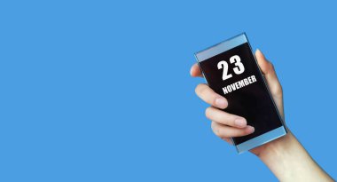 23 Kasım 'da. Ayın 23. günü, takvim tarihi. Kadının elinde mavi izole arka planda boş ekranlı cep telefonu var. Sonbahar ayı, yılın günü konsepti..