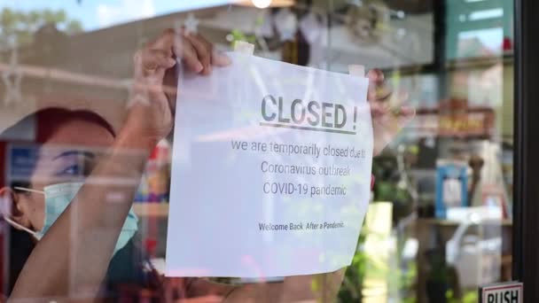 Азійка, одягнена в маску обличчя під час прикріплення бізнесу закритий знак на вході в двері магазину через спалах епідемії коронавірусу covid-19 по всьому світу — стокове відео