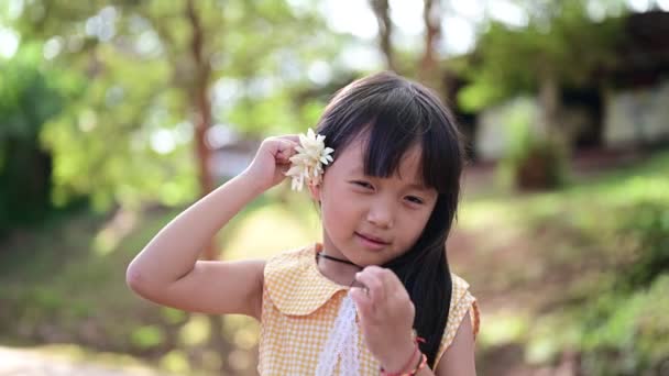 Kulit gadis asia tersenyum gembira dia menggunakan bunga putih untuk menyelipkan telinganya dan melihat kamera sambil tersenyum — Stok Video