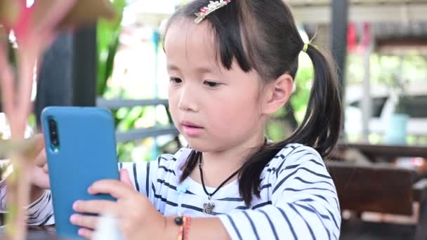 Ázsiai kislány kéz gazdaság és nézni játék smartphone kávézóban. gyerek rabja játékok problémák a gyermekek használja okostelefon technológia Természetes fény kültéri életmód — Stock videók