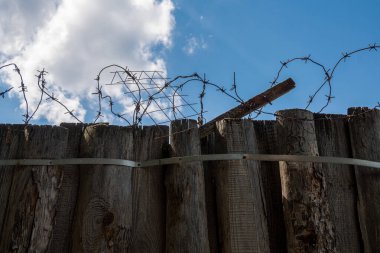 Eski paslı dikenli tel bobinlerin silueti eski, yıpranmış ahşap bir çitin tepesinde parlak mavi bir gökyüzüne ve bulutlara karşı. Boşluğu kopyala.
