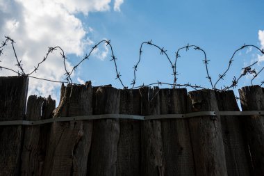 Eski paslı dikenli tel bobinlerin silueti eski, yıpranmış ahşap bir çitin tepesinde parlak mavi bir gökyüzüne ve bulutlara karşı. Boşluğu kopyala.