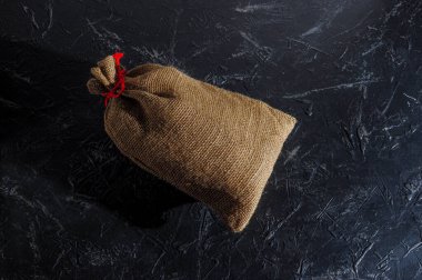 Kaba bir çuval bezinden yapılmış, koyu bir arka planda kırmızı sicimle bağlanmış, mesaj için yer açılmış bir Noel çantası. Boşluğu kopyala.