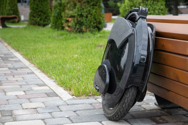 Black electric mono wheel, innovative personal vehicle, self-balancing electric unicycle, ecological urban transport of the future near a wooden bench in a city park.