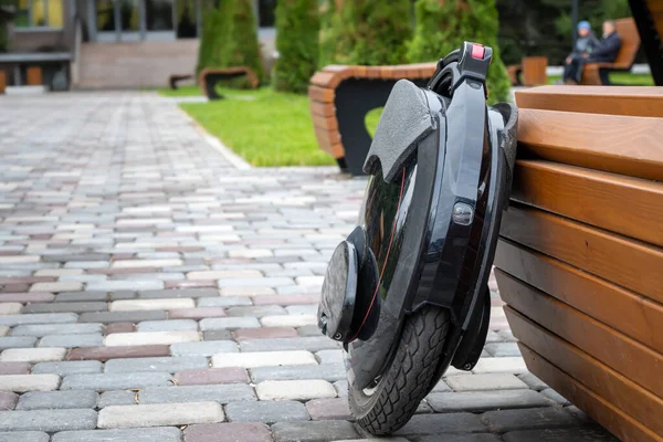 Black electric mono wheel, innovative personal vehicle, self-balancing electric unicycle, ecological urban transport of the future near a wooden bench in a city park.