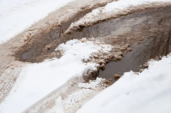 Asfalt yüzeyinde erimiş kar, derin durgun su ve kenarlarında kar. Baharın başında karlı bir yolda su birikintileri.