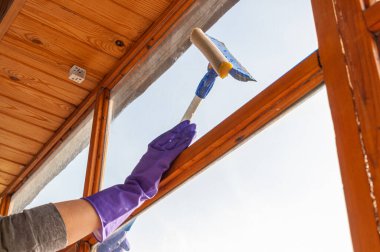 Mavi eldivenli tanınmayan genç bir kadın uzun saplı özel bir kazıyıcıyla balkon camını dışarıdan yıkıyor.
