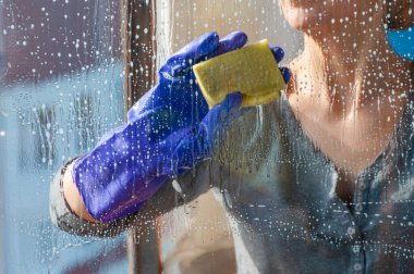Koruyucu mavi eldivenli tanınmayan bir kadın kirli camları süngerle yıkıyor.