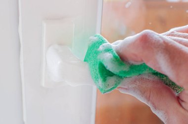 Sabun köpüğü içinde sünger tutan bir adamın yakın çekimi. Beyaz plastik bir kapının sapını yıkayan bir el..