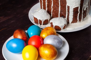Beyaz kremayla kaplanmış ev yapımı Paskalya pastaları ve koyu ahşap arka planda tabakta çok renkli Paskalya yumurtaları..