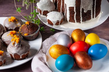 Beyaz kremayla ya da portakal kabuğuyla kaplı ev yapımı Paskalya pastaları. Koyu ahşap arka planda, tabakta renkli Paskalya yumurtaları.