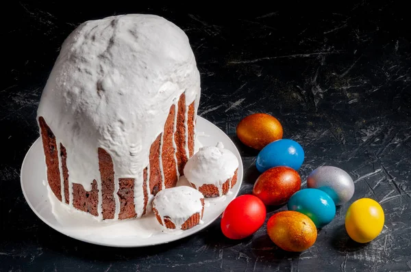 Homemade craft Easter cakes covered with white icing and multicolored painted Easter eggs on plates on a dark background.