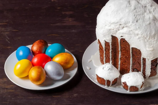 Homemade craft Easter cakes covered with white icing and multicolored painted Easter eggs on plates on a dark wooden background.