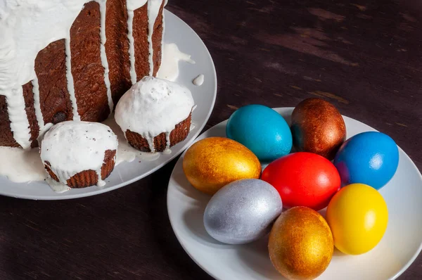 Homemade craft Easter cakes covered with white icing and multicolored painted Easter eggs on plates on a dark wooden background.