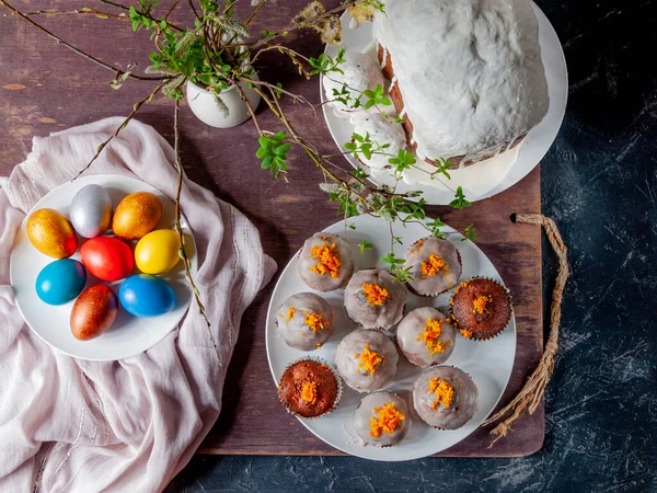 Homemade Easter cakes covered with white icing or orange peel, colorful painted Easter eggs on plates on a dark wooden background