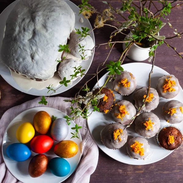 Homemade Easter cakes covered with white icing or orange peel, colorful painted Easter eggs on plates on a dark wooden background