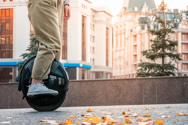 The legs of a young woman ready to ride an electric unicycle, abbreviated EUC, against the background of an evening or morning urban landscape. An unrecognizable woman on an electric monowheel.