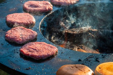 Biftek köfteleri ve hamburger çörekleri açık havada büyük yuvarlak ahşap yakma ızgarasında kızartılıyor, yakın plan. Şehir parkında barbekü festivali. Sokak Fast food.